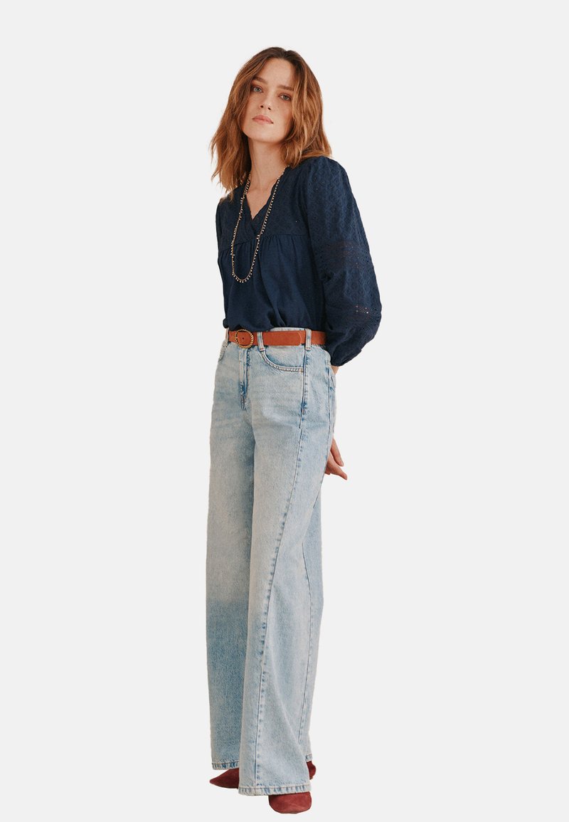 Marineblaue Spitzenbluse mit V-Ausschnitt, kombiniert mit hoch taillierten, hellblauen Jeans im weit geschnittenen Stil und einem braunen Ledergürtel. Rote Schuhe.