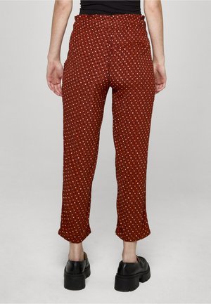 Nő magas derekú rozsdavörös nadrágban, kis fehér és fekete pöttyös mintával, fekete, masszív talpú platform cipővel párosítva, hátulnézetben áll.