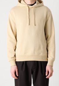 Sudadera beige hecha de tela suave, con cordón, puños y dobladillo acanalados, y un logo discreto en el pecho. Combinada con pantalones oscuros.
