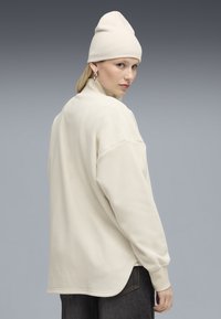 Cremfarbener, oversized Pullover aus strukturiertem Stoff, mit hohem Kragen, überschnittenen Schultern und abgerundetem Saum, kombiniert mit einer passenden Mütze.