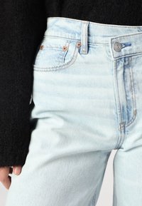 Vaaleansiniset farkut korkealla vyötäröllä, joissa on kuparinväriset napit, klassinen viiden taskun muotoilu ja hieman haalistunut pinta.