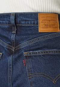 Jeans de mezclilla azul oscuro de cintura alta, con costuras amarillas, un parche de piel marrón en la parte trasera y una etiqueta roja en el bolsillo lateral.