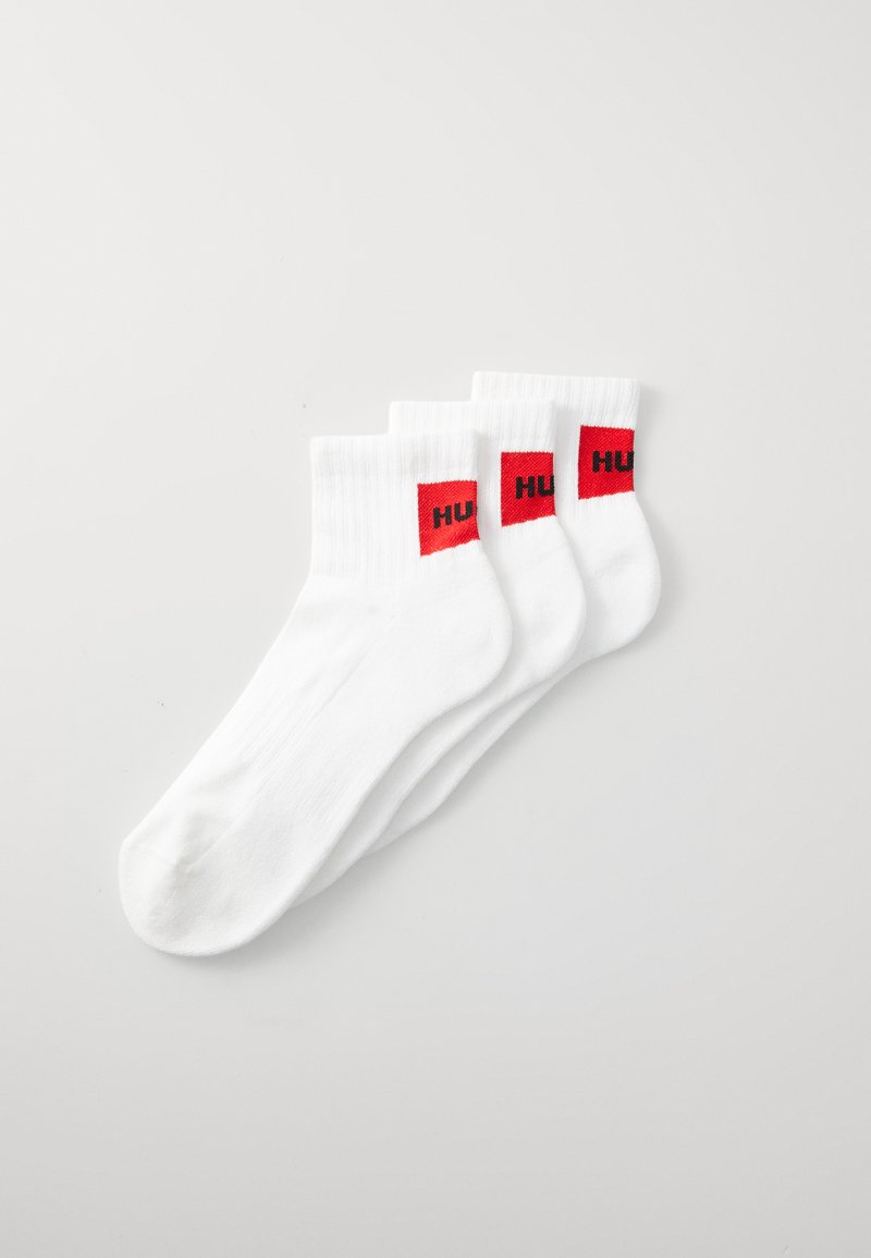 Calcetines de algodón blanco con textura acanalada, que presentan parches de logotipo rectangular rojos etiquetados "HUF" en el tobillo. Se muestran tres pares.