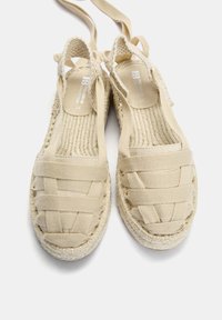Béžové tkané espadrilky s křížovým designem popruhů, plátěným materiálem a jutovou podrážkou, které mají měkkou texturu a plochý podpatek.