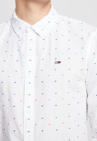 Camicia bianca con bottoni caratterizzata da pois ricamati multicolori in rosa, blu e giallo, con una piccola toppa del marchio sul petto.