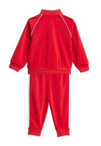 Rød treningsdress laget av glatt stoff, med en glidelåsjakke med høy krage og bukser med hvite sidestriper. Elastiske mansjetter og midjebånd.