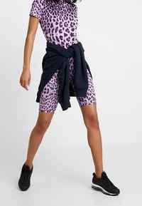 Persona que lleva un mono de manga corta con estampado de leopardo púrpura, zapatillas negras y un sudadera negra atada a la cintura.