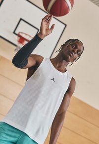 Bílý bezramenný sportovní dres s perforacemi po celé ploše, s černým logem Jumpman. Nosí černý kompresní rukáv a tyrkysové šortky.