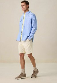 Lichtblauw overhemd met knopen en twee voorkruiden, gele T-shirt eronder, crèmekleurige shorts en beige sportschoenen met grijze accenten.
