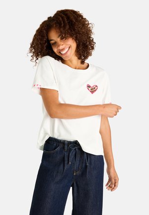 Smiling woman with curly hair wears a white T-shirt with a leopard print heart patch and dark blue denim pants.