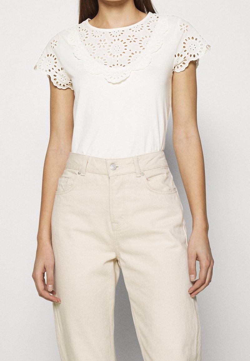 Biały top z koronkowymi zdobieniami wokół dekoltu i krótkimi rękawami, w połączeniu z jasnobeżowymi spodniami z wysokim stanem. Gładka faktura, casualowy design.
