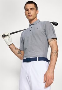 Camisa pólo cinza com botões e logótipo 'CK', combinada com calções brancos. Um cinto azul-marinho apresenta uma fivela 'CK'. Uma mão com luva segura um taco de golfe.