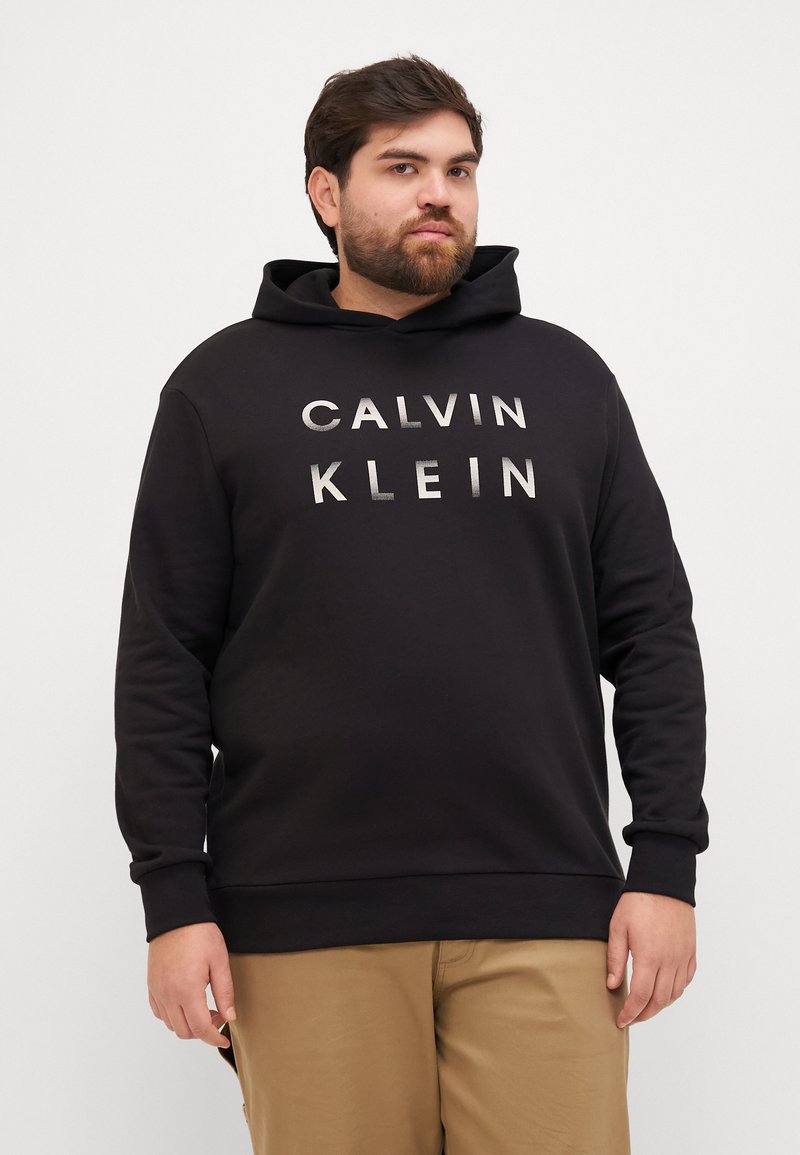 Čierna mikina s kapucňou s potlačou "CALVIN KLEIN" v striebornej farbe, obsahujúca klokanie vrecko a kapucňu so šnúrkou, vyrobená z textúrovaného zmesového bavlneného materiálu.