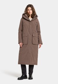 Brauner langer Parka mit Kapuze, der zwei große Fronttaschen, einen Reißverschluss und eine glatte Textur aufweist. Wird über dunklen Hosen und Stiefeln getragen.