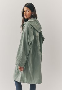 Light green, hooded coat with a textured finish. Features a relaxed fit, dropped shoulders, and side pockets. Long sleeves with button cuffs.