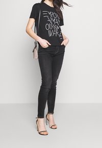 Černé tričko s krátkým rukávem a bílým grafickým textem, kombinované s černými skinny džíny a černými sandály na vysokém podpatku s řemínky. Malá kabelka.