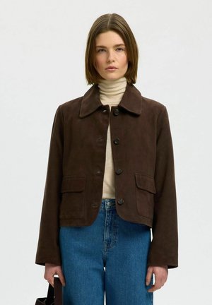 Jeune femme portant une veste marron foncé boutonnée sur un col roulé beige et un jean bleu, tenant un sac à main marron assorti.