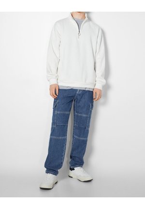 Personne portant un sweat-shirt blanc à fermeture quart zip, un jean cargo bleu avec poches plaquées, et des baskets blanches, se tenant devant un fond blanc uni.