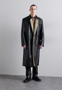 Long black leather coat with oversized lapels and fur lining, paired with fitted black pants and shoes, against a simple background.