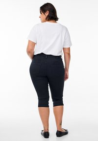 Lühikeste tumedate juustega naine, kes kannab valget t-särki, musta lühikest püksipükse ja musta madalat jalanõud seistes valgel taustal selg vaataja poole.