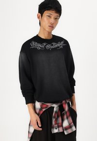 Black sweatshirt with "Eternal Redemption" in silver script across the chest, oversized fit, worn over black pants and red plaid shirt tied at the waist.