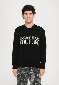 Črn pleten pulover z bold belim napisom "VERSACEJEANS COUTURE" čez prsni del. Okrogla izreza in dolgi rokavi.