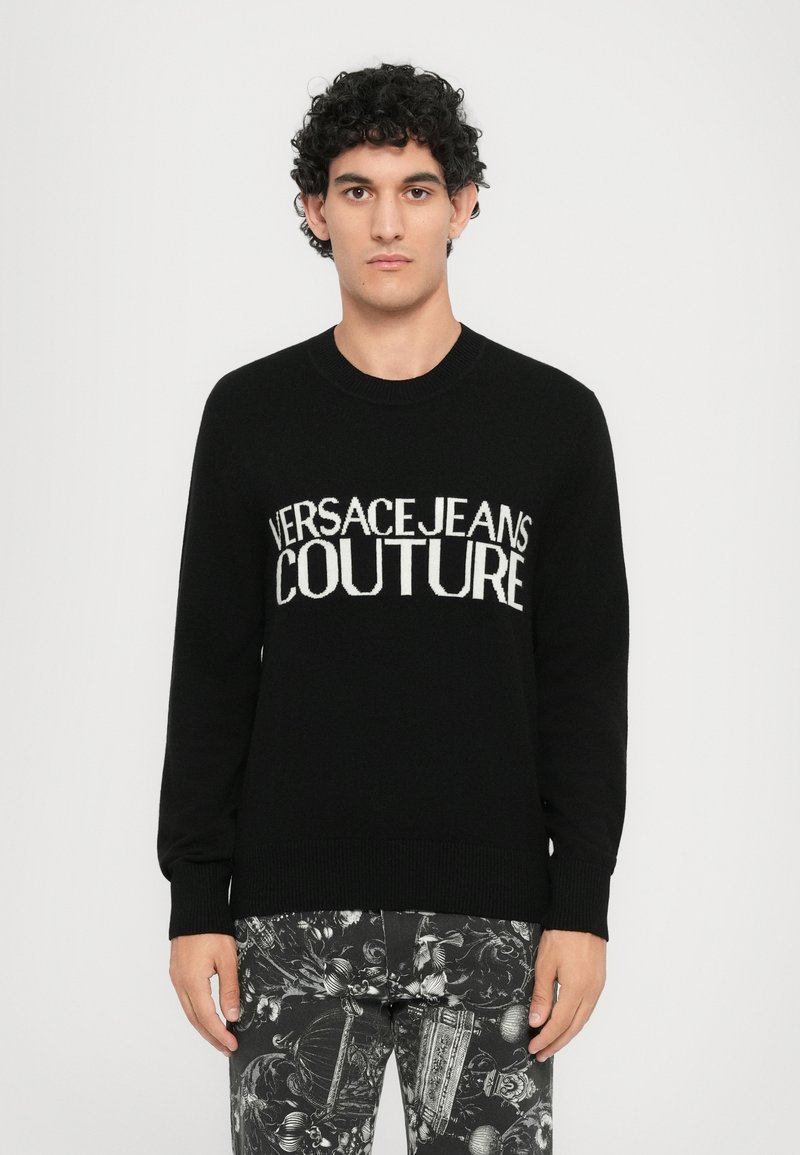 Črn pleten pulover z bold belim napisom "VERSACEJEANS COUTURE" čez prsni del. Okrogla izreza in dolgi rokavi.