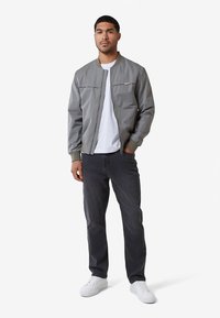 Graue Bomberjacke mit Reißverschluss vorne, gerippten Bündchen und zwei Vorderentaschen, getragen über einem weißen T-Shirt und kombiniert mit dunklen Jeans und weißen Sneakern.