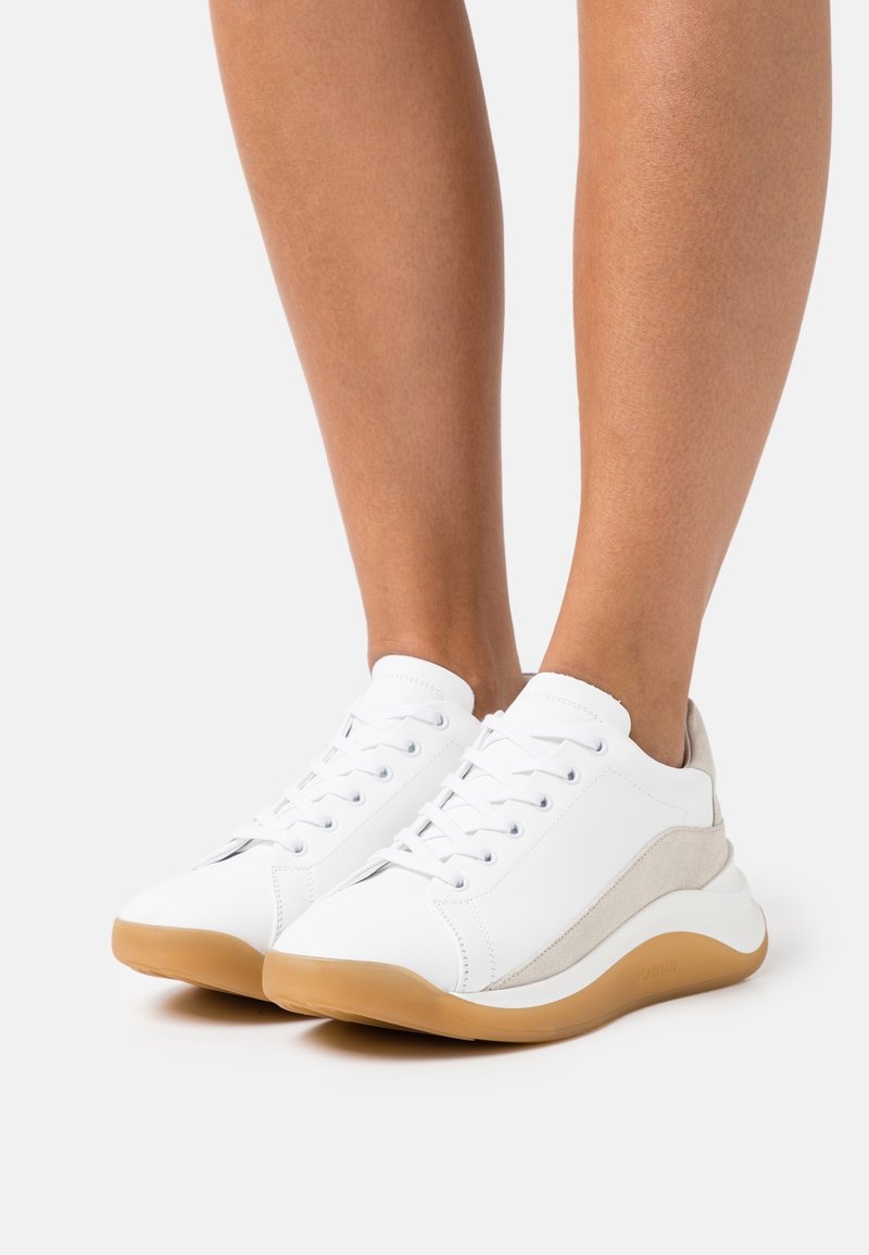 Baskets blanches avec panneaux latéraux en daim beige et semelles en gomme, portées pieds nus sur un fond clair uni.