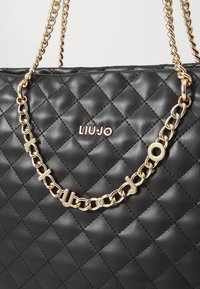 Sac à main matelassé noir avec une bandoulière en chaîne dorée arborant des accents de lettres. Logo "LIU-JO" affiché en évidence. Finition en cuir texturé.