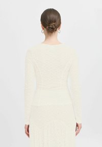 Top ajusté à manches longues en dentelle crème clair avec un motif en losanges texturé. Tissu lisse à la taille, col rond. Vue de dos.