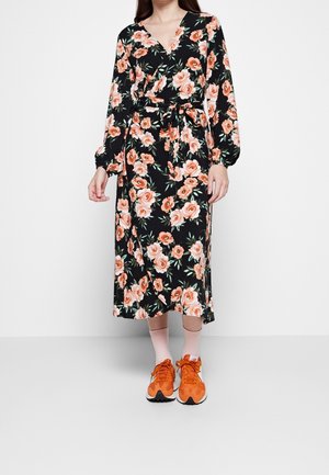 Kvinde iført en sort blomsterprint slå-om kjole med lange ærmer og orange sneakers med lyserøde sokker, stående foran en hvid baggrund.