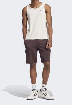 Mies, jolla on päällä valkoinen Adidas-urheilotoppi, tummat ruskeat cargo-shortsit, valkoiset Adidas-sukat ja valkoiset Adidas-tennarit yksinkertaista taustaa vasten.