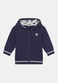 Felpa con zip blu navy con logo bianco, polsini a righe e fodera interna a motivi con varie illustrazioni grafiche.