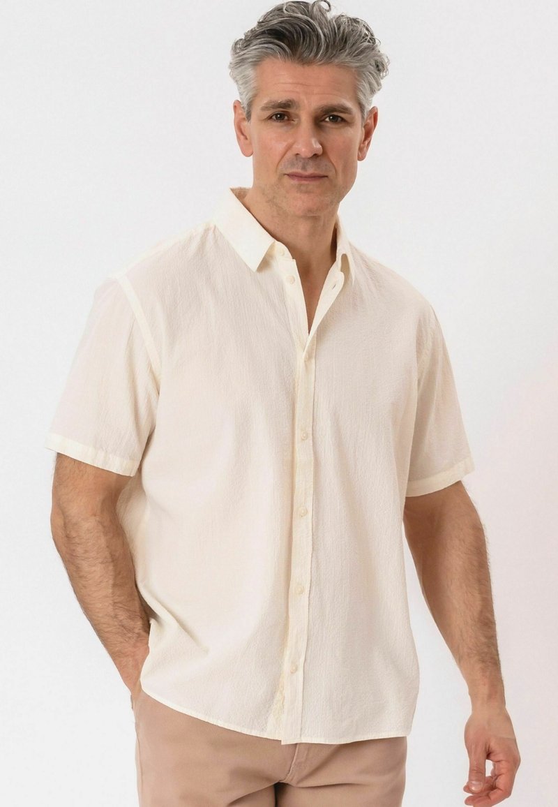 Homme d'âge moyen aux cheveux gris portant une chemise crème à manches courtes avec boutons et un pantalon marron clair, debout devant un fond uni.