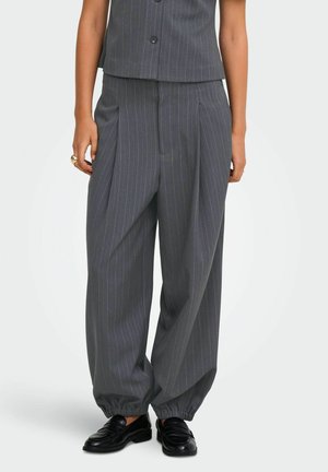 Pantalon ample gris foncé à fines rayures avec poignets élastiques, porté avec des mocassins noirs et un gilet sans manches assorti et boutonné.