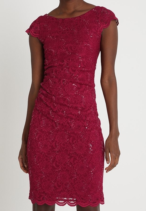 Robe en dentelle bordeaux ajustée, longueur genou, avec motif floral et manches courtes, portée par une personne debout devant un fond uni.