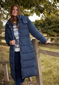 Abrigo de plumas largo en azul marino con acolchado horizontal, con un cuello alto, cierre frontal con cremallera y bolsillos laterales, combinado con un suéter estampado.
