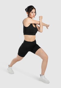 Top sportivo nero e pantaloni corti a vita alta realizzati in tessuto elastico. Il modello si sta stiracchiando indossando scarpe da ginnastica bianche. Design semplice e aderente.