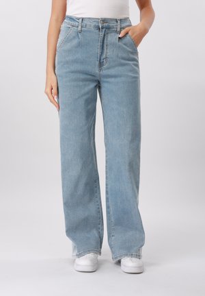 Osoba nosiaca svetlomodré široké rifle a biele tenisky, s jednou rukou v vrecku a viditeľným jednoduchým bielym topom.