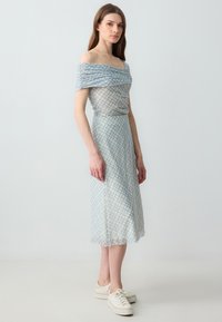 Off-shoulder, lichtblauwe jurk met een geruit patroon, gerimpelde bodice en vloeiende rok, gecombineerd met witte sneakers.