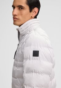 Helle graue Steppjacke mit hohem Kragen, gestepptem Textil und einem schwarzen Logopatch auf der linken Schulter. Frontreißverschluss.