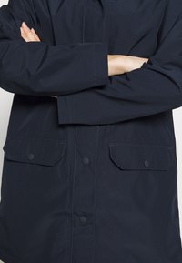 Personne portant une veste bleu marine foncé avec des poches à rabat boutonnées, bras croisés sur la poitrine, visible du cou jusqu’aux hanches.