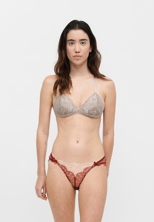 Žena, ktorá má na sebe béžovú čipkovanú podprsenku a červené čipkované nohavičky s mašličkovými detailmi, stojaca pred jednofarebným bielym pozadím, tvárou vpred.