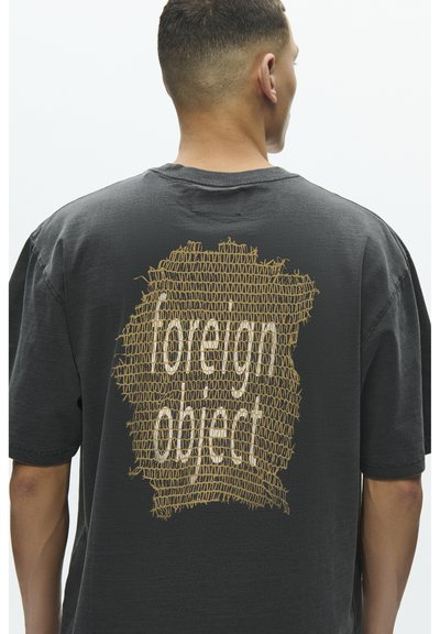 Svart oversized t-skjorte med en strukturert gyllen grafikk på baksiden med teksten "foreign object" i et unikt mønster. Laget av bomull.