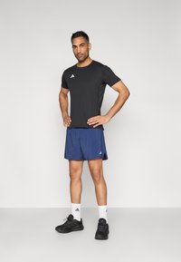 Svart trenings-T-skjorte med hvit logo, blå shorts, hvite sokker og svarte joggesko. Modell står med hendene på hoftene.