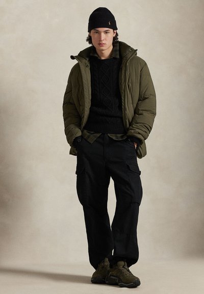 Casaco acolchoado verde-oliva, sweater de malha grossa preto e calças cargo pretas. Gorro escuro e ténis desportivos verdes completam o conjunto.