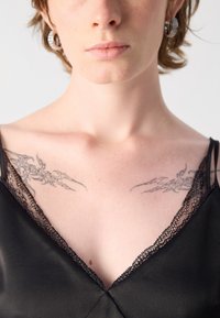 Schwarzes Satin-Oberteil mit V-Ausschnitt, verziert mit zarter schwarzer Spitzenbordüre. Auf der Schlüsselbeinregion sind zwei kunstvolle Tattoos sichtbar.