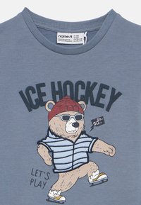 Γκρι βαμβακερό μπλουζάκι με καρτούν αρκούδα με γυαλιά ηλίου και κόκκινο μπερέ, που σκιτάρει σε ριγέ μπλούζα. Το κείμενο γράφει "ΠΑΓΟΧΡΙΣΤΟ" και "ΑΣ ΠΑΙΞΟΥΜΕ".