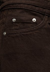 Κοντινή λήψη σκουρόχρωμου καφέ υφάσματος τζιν που δείχνει μια μικρή μπροστινή τσέπη με τρεις ασημένιες στροφίες και ορατές λεπτομέρειες ραφής.